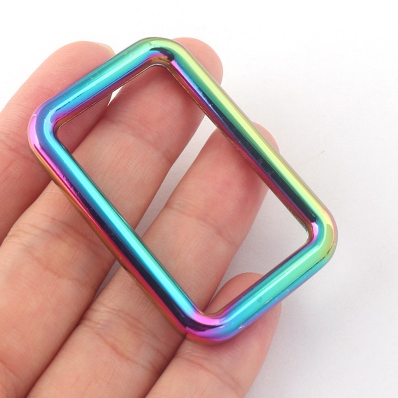 K&ouml;nnte beinhalten: Ein regenbogenfarbener D-Ring aus Metall mit glatter, polierter Oberfl&auml;che. Der Ring ist rechteckig und hat abgerundete Kanten.
