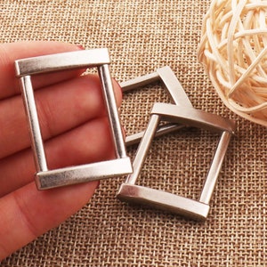 Puede incluir: Tres deslizadores de hebilla de cinturón de metal plateado. Los deslizadores son rectangulares con esquinas redondeadas y tienen un acabado liso y pulido.