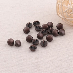 May include: A collection of 17 small, round, bronze-colored buttons with a loop on the back. The buttons are scattered on a light beige fabric surface.