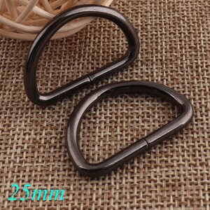 May include: Two black metal D-rings, each measuring 25mm in diameter. The rings are lying on a woven brown fabric background.