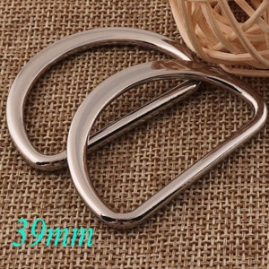 May include: Two silver metal D-ring buckles, each measuring 39mm in diameter. The buckles are stacked on top of each other, with the top buckle slightly offset to the right. The buckles are on a brown woven fabric background.