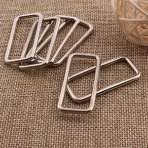 May include: A set of eight silver metal rectangular rings. The rings are stacked on top of each other and are lying on a brown woven fabric surface.