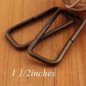 Puede incluir: Dos anillos en D de metal de color dorado antiguo, cada uno mide 3,8 cm de largo. Los anillos están apilados uno encima del otro sobre una superficie de madera.