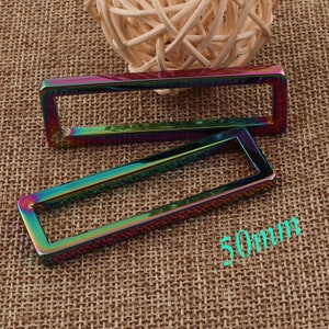 Peut inclure: Deux anneaux rectangulaires en métal iridescent, chacun mesurant 50 mm. Les anneaux sont sur un fond de tissu brun tissé.