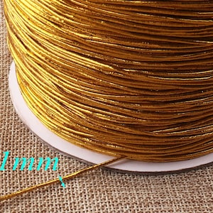 Puede incluir: Una bobina de cordón elástico dorado, de 1 mm de grosor. El cordón está enrollado firmemente alrededor de la bobina, y el extremo del cordón es visible en la esquina inferior izquierda de la imagen.