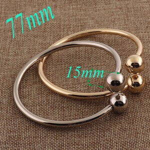 Puede incluir: Se muestran dos brazaletes de metal, uno dorado y otro plateado. Cada brazalete tiene un diseño redondeado con una bola en cada extremo. El brazalete más grande tiene 7,7 cm de diámetro y la bola tiene 1,5 cm de diámetro.