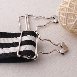 May include: A black and white striped elastic suspender with a silver metal clip. The clip has a heart-shaped design.