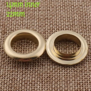 Könnte beinhalten: Zwei goldene Metallösen mit einem Innendurchmesser von 14 mm und einem Außendurchmesser von 22 mm. Die Ösen befinden sich auf einem braunen gewebten Stoffhintergrund.