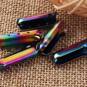 May include: Several iridescent metal shoelace aglets are arranged on a textured surface. The aglets have a rainbow sheen, with colors shifting from blue to green to yellow. Each aglet is a small, rectangular tube with a rounded end and a slot.