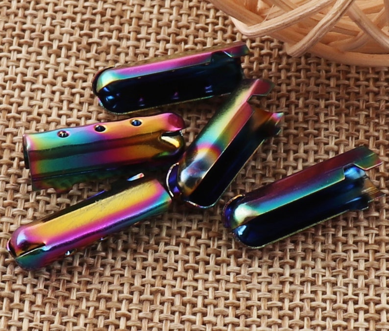 May include: Several iridescent metal shoelace aglets are arranged on a textured surface. The aglets have a rainbow sheen, with colors shifting from blue to green to yellow. Each aglet is a small, rectangular tube with a rounded end and a slot.