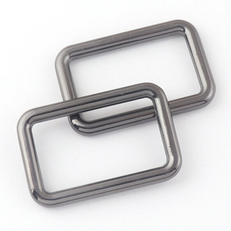 May include: Two black metal D-rings, stacked on top of each other. The rings are rectangular in shape with rounded corners.