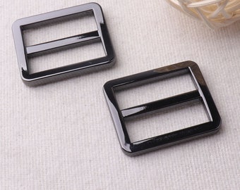 May include: Two black metal slider buckles. The buckles are rectangular with rounded corners and have two horizontal bars.