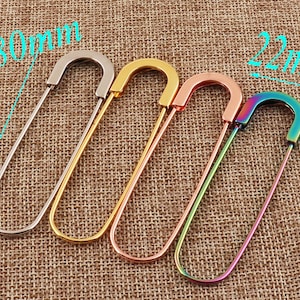 May include: Four safety pins in different colours: silver, gold, rose gold, and iridescent. The pins are 80 mm long and 22 mm wide.