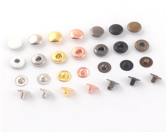 Puede incluir: Surtido de botones a presión metálicos en varios colores y acabados, incluyendo plata, oro, negro y cobre. Los botones a presión están dispuestos en un patrón aleatorio sobre una superficie blanca.