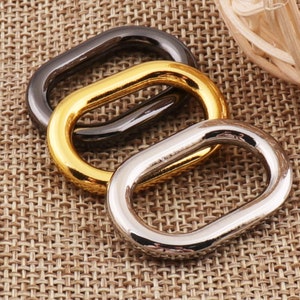 May include: Three oval-shaped metal links in silver, gold, and dark gray. The links are arranged on a textured, tan surface. The links are likely used for jewelry making or crafting.