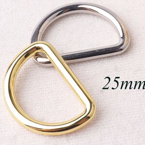 May include: Two metal D-rings, one gold and one silver, with a 25mm diameter. The rings are lying on a white fabric surface.