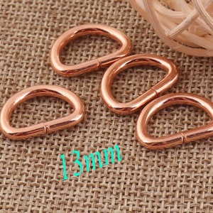 May include: Four rose gold metal D-rings, each measuring 13 mm in diameter, are arranged on a woven brown fabric background.