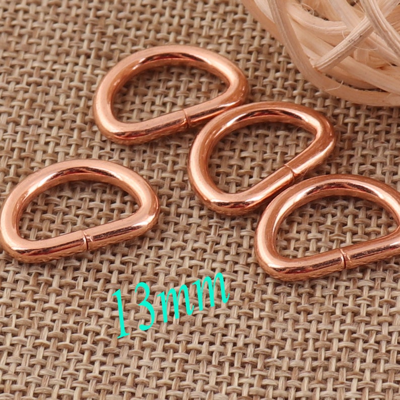 May include: Four rose gold metal D-rings, each measuring 13 mm in diameter, are arranged on a woven brown fabric background.