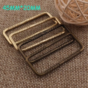 May include: Three rectangular metal buckles in gold and bronze tones. The buckles, measuring 4.5cm x 2cm, feature a textured pattern. The background is a woven fabric.