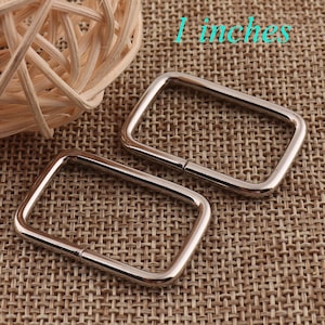 May include: Two silver-toned metal D-rings, each measuring 1 inch in length, are displayed on a brown woven fabric background. The D-rings are rectangular in shape with rounded corners.