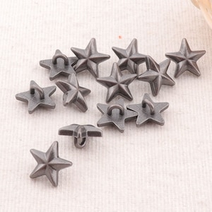 Puede incluir: Un conjunto de 12 botones de metal en forma de estrella de color plateado con un bucle en la parte posterior. Los botones están esparcidos sobre una superficie de tela gris claro.