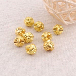 Puede incluir: Diez botones de metal de color dorado con un diseño de nudo tejido. Los botones están esparcidos sobre una superficie de tela beige claro.