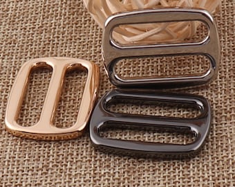 May include: Three metal slider buckles in different finishes: gold, black, and silver. The buckles are rectangular with two parallel slots.