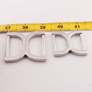May include: Two silver metal D-ring buckles, one measuring 3.75 inches and the other measuring 2.75 inches, are shown next to a yellow measuring tape.