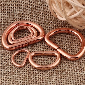 May include: Four rose gold metal D-rings of different sizes. The rings are arranged on a brown woven fabric background.