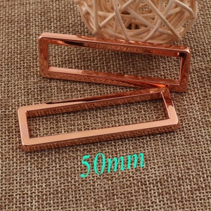 May include: Two rose gold metal rectangular frames, each measuring 50mm in length. The frames are on a brown woven fabric background.