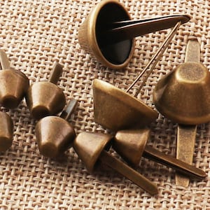 May include: A collection of antique gold-toned metal rivets with pointed tips. The rivets have a cone-shaped head and are designed for attaching fabric or leather.