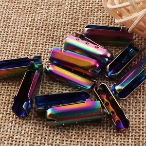 May include: A collection of iridescent metal cord ends. The cord ends are a mix of blues, greens, and purples, with a shiny, metallic finish. They have a rectangular shape with a curved top and small holes for attaching to cords or other materials.