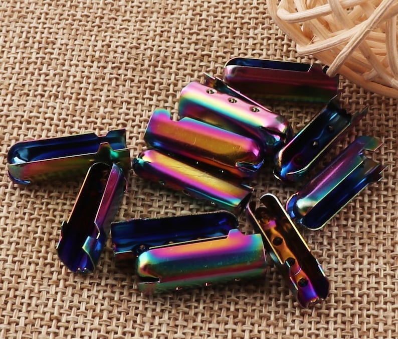 May include: A collection of iridescent metal cord ends. The cord ends are a mix of blues, greens, and purples, with a shiny, metallic finish. They have a rectangular shape with a curved top and small holes for attaching to cords or other materials.