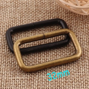 May include: Two metal D-rings, one black and one gold, on a brown woven fabric background. The gold D-ring is 32mm in size.