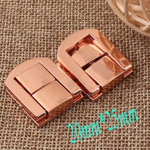 Pode incluir: Dois fechos de metal cor de ouro rosa, cada um com forma retangular e um design de dobradiça. Os fechos medem 20 mm por 25 mm.