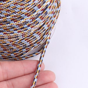 Op de afbeelding: Een close-up van een veelkleurig gevlochten koord met gele, rode, blauwe en witte strepen. Het koord is om een spoel gewikkeld en een lengte van het koord is uitgerekt.