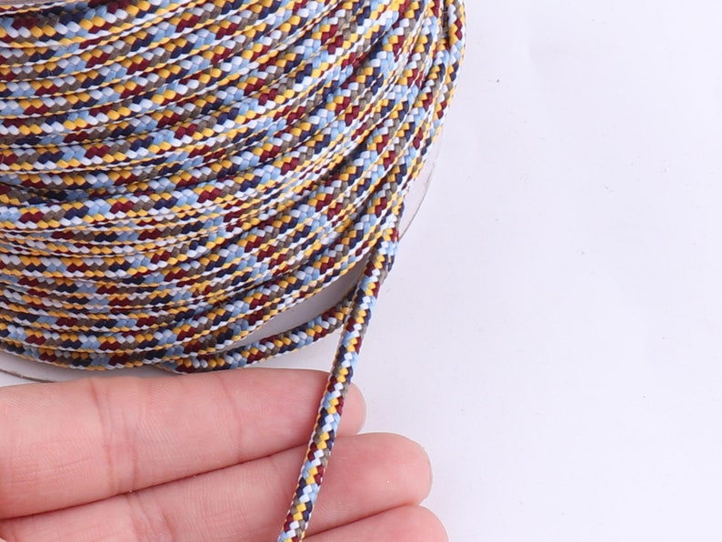 Op de afbeelding: Een close-up van een veelkleurig gevlochten koord met gele, rode, blauwe en witte strepen. Het koord is om een spoel gewikkeld en een lengte van het koord is uitgerekt.