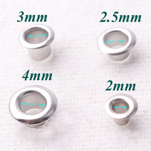 Peut inclure: Quatre œillets en métal argenté de différentes tailles : 2 mm, 2,5 mm, 3 mm et 4 mm. Les œillets sont présentés sur un fond en tissu blanc.