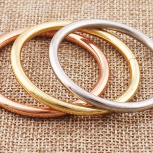 May include: Three interlocking metal rings, one silver, one gold, and one rose gold, on a textured brown surface.