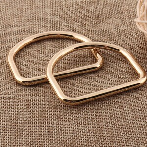 May include: Two gold-toned metal D-rings, one slightly larger than the other, are shown on a brown textured surface. The rings are smooth and have a polished finish.