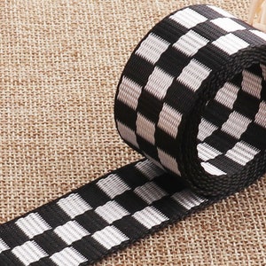 May include: Black and white checkered ribbon with a woven texture. The ribbon is rolled up and the checkered pattern is visible on both the top and side of the roll.