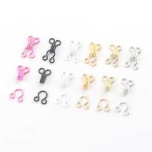 May include: Assortment of metal hook and eye closures in various colors, including pink, black, white, gold, and silver. These small fasteners are arranged on a white surface, showcasing their different finishes and designs. Ideal for sewing and crafting projects.