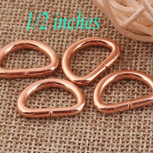 May include: Four rose gold D-rings, each measuring 1/2 inch in diameter. The rings are on a woven brown fabric background.
