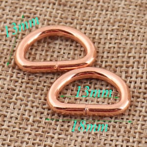 May include: Two 13mm rose gold D-rings, each measuring 18mm in length. The rings are shown on a woven brown fabric background.