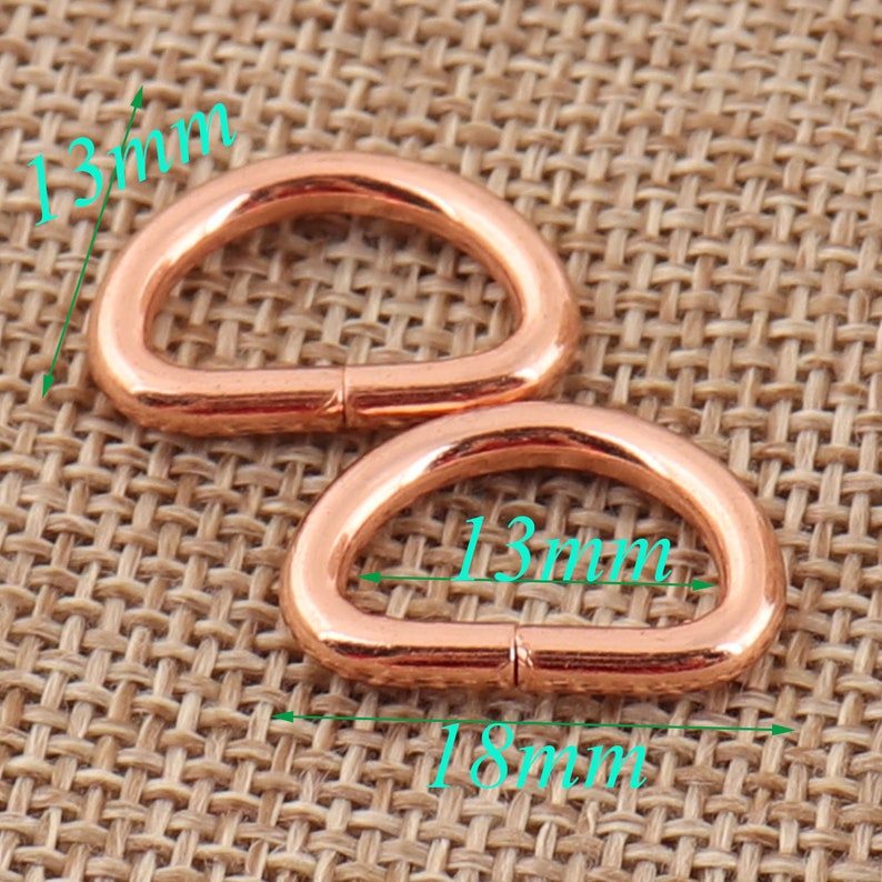 May include: Two 13mm rose gold D-rings, each measuring 18mm in length. The rings are shown on a woven brown fabric background.