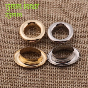Op de afbeelding: Vier metalen oogjes, twee goud en twee zilver, met een binnendiameter van 15 mm en een buitendiameter van 23 mm. De oogjes zijn in een vierkant patroon op een bruine stoffen achtergrond gerangschikt.