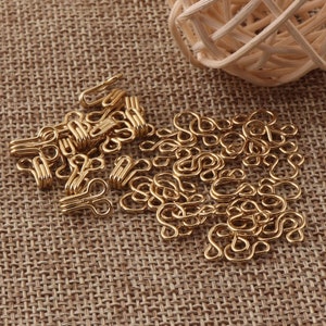 May include: A pile of gold-colored metal hook and eye closures. The hooks and eyes are arranged in a random pattern on a brown burlap surface.