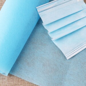 May include: A roll of light blue non-woven fabric, a type of material often used for crafting and DIY projects, is shown next to a folded light blue surgical mask.