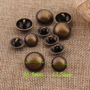 Può includere: Un gruppo di 10 bottoni in metallo color oro antico con forma a cupola rialzata e un piccolo anello sul retro. I bottoni hanno un diametro di 4,2 cm e 3,2 cm.
