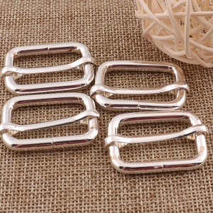 May include: Four silver metal slider buckles, each with a rectangular shape and two loops. The buckles are arranged in a row on a brown woven fabric background.